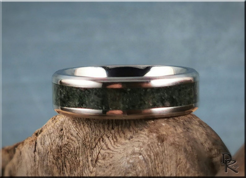 K&ouml;nnte beinhalten: Ein silberner Ring mit breitem Band und einem dunkelgr&uuml;nen, strukturierten Einsatz. Das polierte Metall des Rings kontrastiert mit dem nat&uuml;rlichen, steinartigen Aussehen des Einsatzes. Der Ring ist vor einem neutralen Hintergrund ausgestellt.
