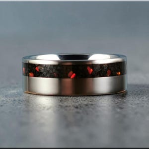 May include: A silver ring with a black center band speckled with red accents. The ring has a polished, metallic finish and a modern design. The band is approximately 6 mm wide.