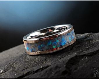 May include: A silver ring with a band of blue and green opal-like material. The ring has a polished silver interior and a gold-coloured border. The ring is set on a dark, textured surface.