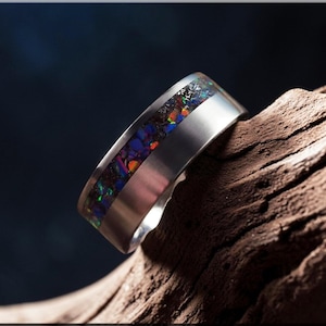 May include: A silver ring with a band of colorful, iridescent opal. The opal features shades of blue, purple, orange, and green. The ring is set against a dark background and rests on a piece of wood.