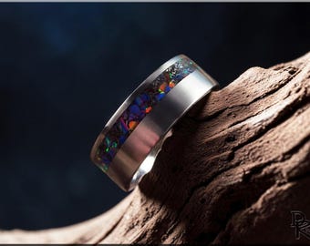 May include: A silver ring with a band of colourful, iridescent opal. The opal features shades of blue, purple, orange, and green. The ring is set against a dark background and rests on a piece of wood.