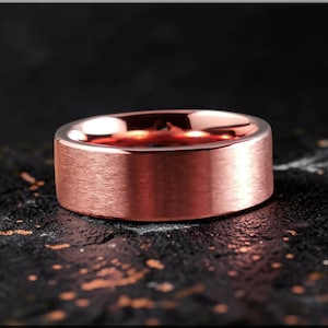 Op de afbeelding: Een roségouden ring met een geborstelde metalen afwerking. De ring heeft een platte, brede band. De binnenkant van de ring reflecteert een roodachtige tint. De ring wordt gepresenteerd op een donker, getextureerd oppervlak.