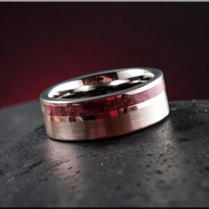 May include: A silver ring with a brushed metal finish and a band of deep red stone. The ring is displayed against a dark, textured surface, showcasing its polished details. A piece of jewellery.