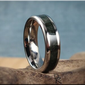 K&ouml;nnte beinhalten: Ein polierter Silberring mit einer Einlage aus dunkelgr&uuml;nem Stein. Der Ring hat eine glatte, reflektierende Oberfl&auml;che und ein Band aus dunkelgr&uuml;nem Stein. Der Ring wird auf einem St&uuml;ck Treibholz vor einem verschwommenen blauen Hintergrund pr&auml;sentiert.