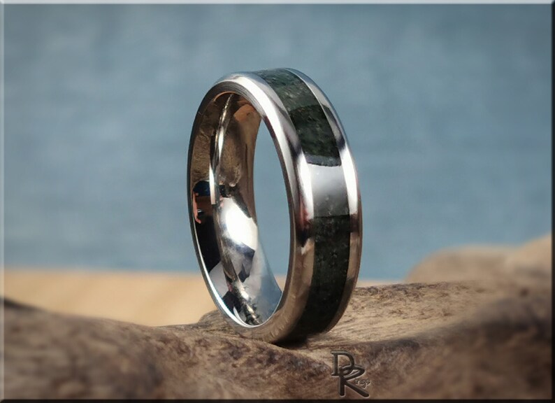 K&ouml;nnte beinhalten: Ein polierter Silberring mit einer Einlage aus dunkelgr&uuml;nem Stein. Der Ring hat eine glatte, reflektierende Oberfl&auml;che und ein Band aus dunkelgr&uuml;nem Stein. Der Ring wird auf einem St&uuml;ck Treibholz vor einem verschwommenen blauen Hintergrund pr&auml;sentiert.