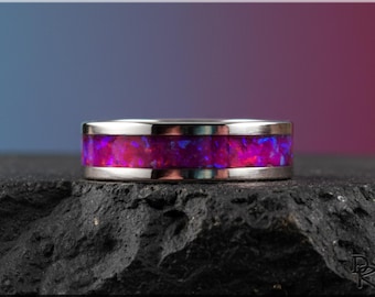 May include: A silver ring with a vibrant, multi-coloured opal inlay. The opal features shades of pink, purple, and blue. The ring is set against a dark, textured surface with a gradient background of blue and red.