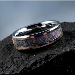 Puede incluir: Un anillo plateado con una banda de piedra rosa y gris, enmarcada por oro rosa y plata. El anillo se muestra sobre una superficie oscura y texturizada, con un fondo borroso.