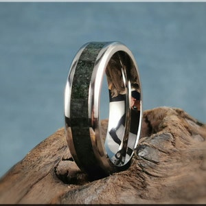 K&ouml;nnte beinhalten: Nahaufnahme eines silbernen Rings mit einer dunkelgr&uuml;nen Steineinlage. Der Ring ist hochglanzpoliert und spiegelt die Umgebung wider. Der Ring liegt auf einem St&uuml;ck Treibholz.