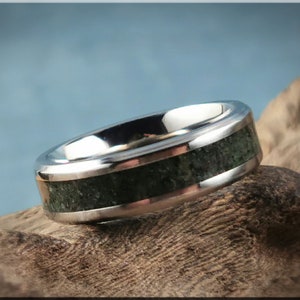 K&ouml;nnte beinhalten: Ein silberner Ring mit einem dunkelgr&uuml;nen Steineinsatz. Der Ring hat eine polierte, reflektierende Oberfl&auml;che und ein Band aus dunkelgr&uuml;nem, strukturiertem Material. Der Ring wird auf einem St&uuml;ck Holz pr&auml;sentiert.