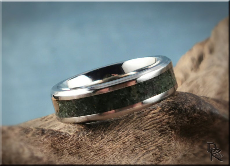 K&ouml;nnte beinhalten: Ein silberner Ring mit einem dunkelgr&uuml;nen Steineinsatz. Der Ring hat eine polierte, reflektierende Oberfl&auml;che und ein Band aus dunkelgr&uuml;nem, strukturiertem Material. Der Ring wird auf einem St&uuml;ck Holz pr&auml;sentiert.