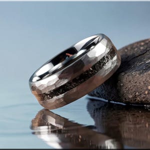 May include: A silver ring with a hammered texture and a dark, textured inlay. The ring is resting on a dark, rough stone, with a reflective surface below. The ring's design is modern and eye-catching.