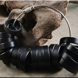 May include: A black ring sizer tool with multiple ring sizes, connected by a silver cable. The ring sizes are marked with white numbers. The tool is displayed on a wooden background.