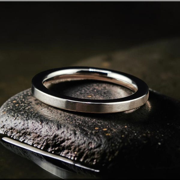 May include: A silver band ring with a flat profile rests on a dark, textured stone. The ring's polished surface reflects light, highlighting its smooth, minimalist design. The ring is a simple, elegant piece of jewellery.