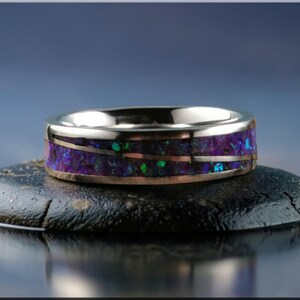 May include: A silver ring with a band of purple, green, and copper-colored inlay, resting on a dark, textured stone. The ring's design features a polished silver exterior and a vibrant, colorful interior.