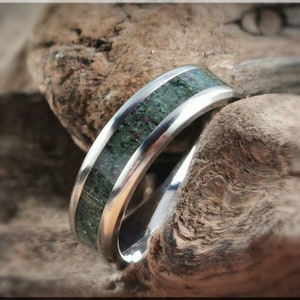 K&ouml;nnte beinhalten: Ein silberner Ring mit einer gr&uuml;nen und roten Steineinlage. Der Ring ist vor einem strukturierten, braunen Holzuntergrund platziert, was den Kontrast zwischen Metall und Naturmaterial hervorhebt.