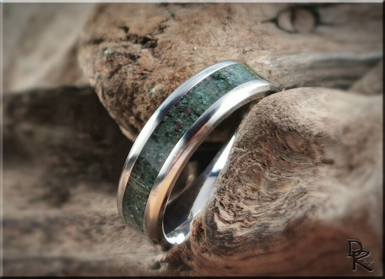 K&ouml;nnte beinhalten: Ein silberner Ring mit einer gr&uuml;nen und roten Steineinlage. Der Ring ist vor einem strukturierten, braunen Holzuntergrund platziert, was den Kontrast zwischen Metall und Naturmaterial hervorhebt.