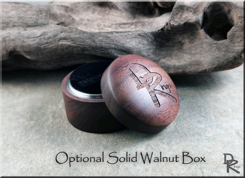 K&ouml;nnte beinhalten: Ge&ouml;ffnete Ringbox aus massivem Walnussholz mit schwarzem Samtinnenraum. Der Deckel ist mit einem Logo graviert und der Text "Optional Solid Walnut Box" befindet sich unter der Box. Die Box steht auf einer grauen Oberfl&auml;che.