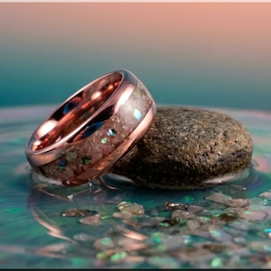May include: A rose gold ring with a unique design rests on a small, textured stone. The ring features a band with a colourful, iridescent inlay. The background is a gradient of teal and peach, with scattered iridescent fragments.