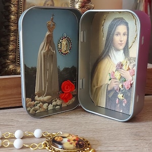 May include: A religious-themed tin box with two compartments, each featuring a printed image. One side depicts a figure in a white robe, the other a woman holding flowers. A rosary with white beads and a gold-colored pendant is in the foreground.