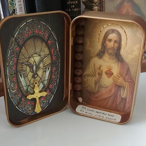 May include: Open, rectangular, gold-coloured tin with two compartments. One side features a stained-glass design with a dove and a gold cross. The other side displays an image of Jesus with a rosary. Text in Latin is visible.