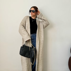 May include: A long, beige cardigan with a hood and a black quilted chain bag. The cardigan is open and the person is wearing blue jeans and black sunglasses.