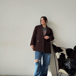 May include: A woman wearing a brown double-breasted blazer, blue jeans with distressed details, and white trainers. She is standing in front of a white wall with a cowhide chair in the background.