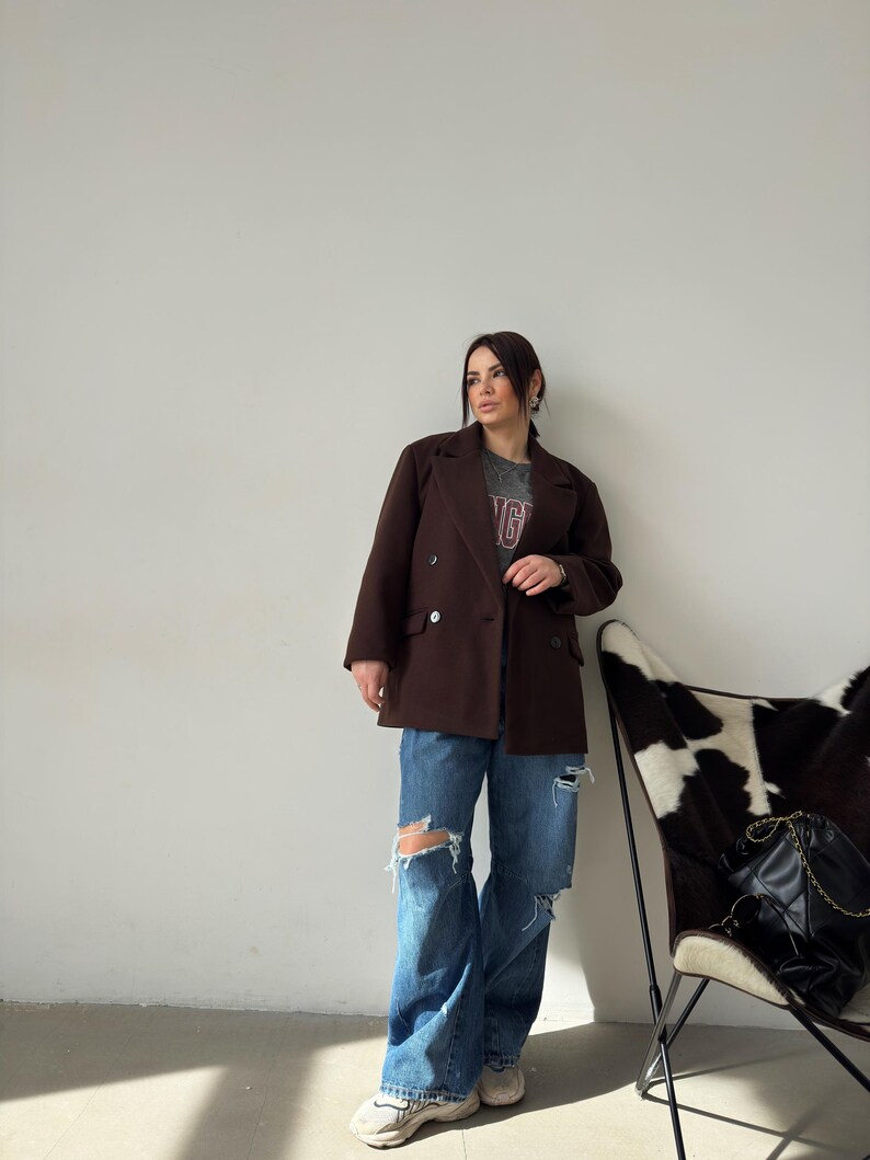May include: A woman wearing a brown double-breasted blazer, blue jeans with distressed details, and white trainers. She is standing in front of a white wall with a cowhide chair in the background.