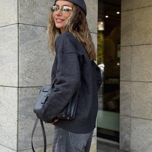 Puede incluir: Una mujer con boina gris oscuro, gafas de sol, suéter y vaqueros. Lleva un bolso negro. El conjunto es monocromático, con énfasis en las texturas y tonos de gris.