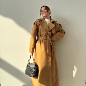 May include: A woman wearing a long camel-colored coat with a belt, a white turtleneck sweater, and blue jeans. She is holding a black handbag with a gold chain strap.