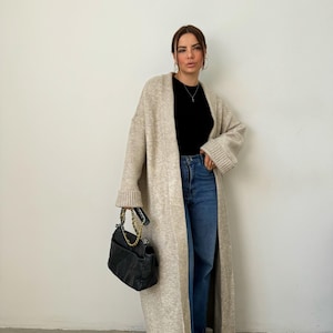 May include: A woman wearing a long, beige cardigan with a black top and blue jeans. She is holding a black handbag with a gold chain strap.
