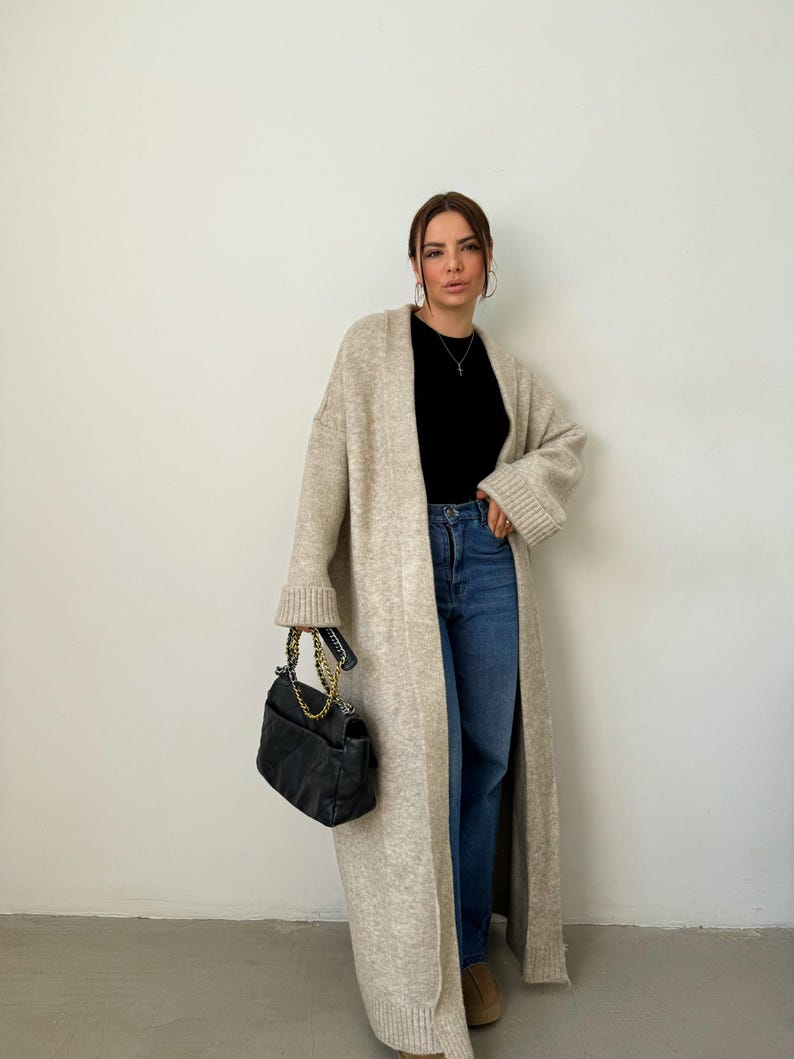 May include: A woman wearing a long, beige cardigan with a black top and blue jeans. She is holding a black handbag with a gold chain strap.