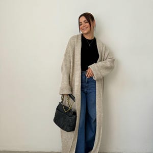May include: A long, beige cardigan with a black top and blue jeans. The cardigan has long sleeves and a relaxed fit. The person is holding a black handbag with a gold chain strap.