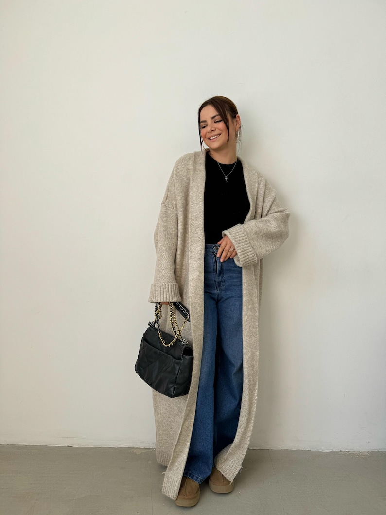 May include: A long, beige cardigan with a black top and blue jeans. The cardigan has long sleeves and a relaxed fit. The person is holding a black handbag with a gold chain strap.
