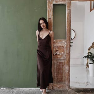 K&ouml;nnte beinhalten: Eine Frau in einem braunen Satin-Schlafkleid mit V-Ausschnitt und d&uuml;nnen Tr&auml;gern. Sie steht vor einer Holzt&uuml;r mit einer gr&uuml;nen Wand dahinter.