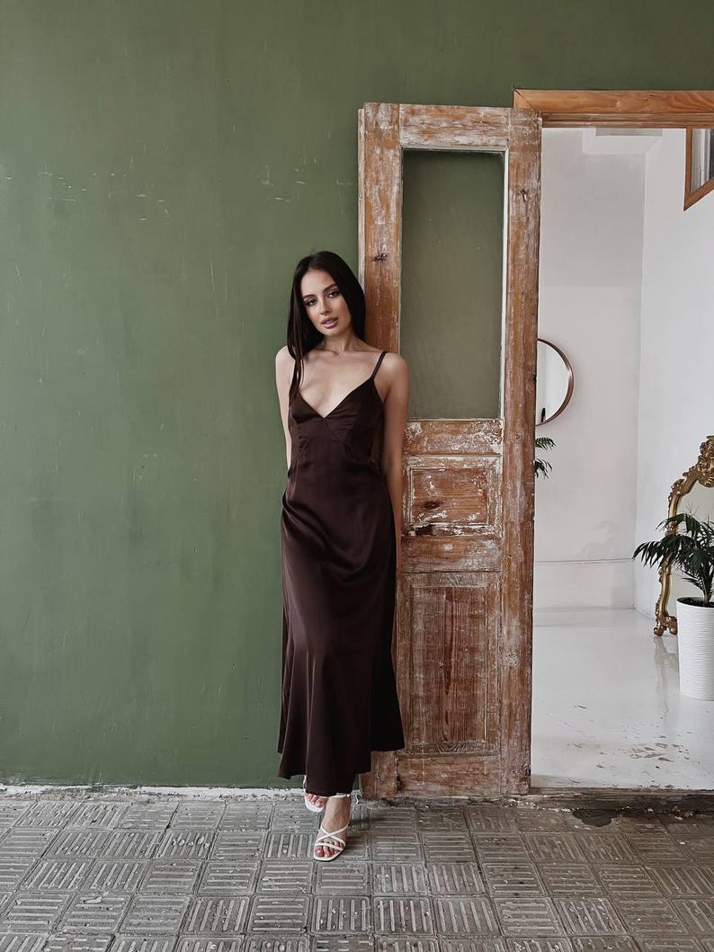K&ouml;nnte beinhalten: Eine Frau in einem braunen Satin-Schlafkleid mit V-Ausschnitt und d&uuml;nnen Tr&auml;gern. Sie steht vor einer Holzt&uuml;r mit einer gr&uuml;nen Wand dahinter.