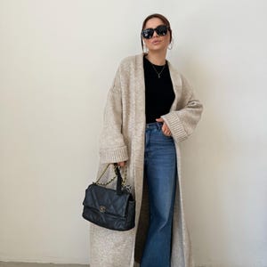 May include: A long, beige cardigan with a black top and blue jeans. The cardigan has long sleeves and a relaxed fit. The person is wearing black sunglasses and a small black handbag.