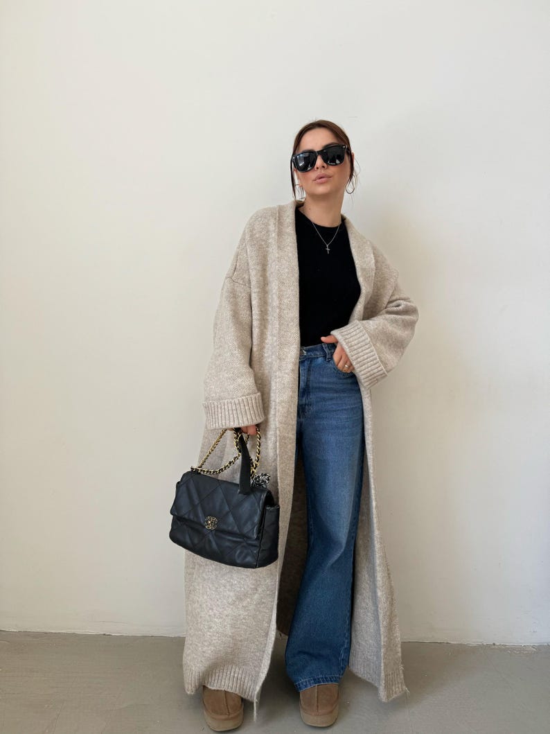 May include: A long, beige cardigan with a black top and blue jeans. The cardigan has long sleeves and a relaxed fit. The person is wearing black sunglasses and a small black handbag.