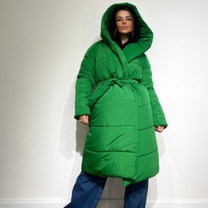 Può includere: Un lungo piumino verde brillante con un ampio cappuccio e una cintura annodata. Il cappotto è indossato sopra una felpa con cappuccio nera e jeans blu. Il cappotto ha un design trapuntato.