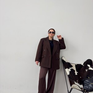 May include: A woman wearing a brown double-breasted blazer and matching wide-leg trousers. She is wearing sunglasses and a black top. She is standing in front of a white wall with a cowhide chair in the background.