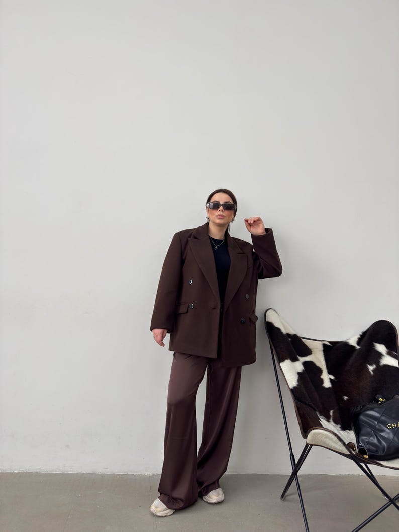 May include: A woman wearing a brown double-breasted blazer and matching wide-leg trousers. She is wearing sunglasses and a black top. She is standing in front of a white wall with a cowhide chair in the background.