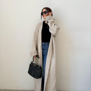May include: A long, beige cardigan with a shawl collar and long sleeves. The cardigan is open in the front and is worn over a black top and blue jeans. The person is wearing black sunglasses and carrying a black handbag with a gold chain strap.