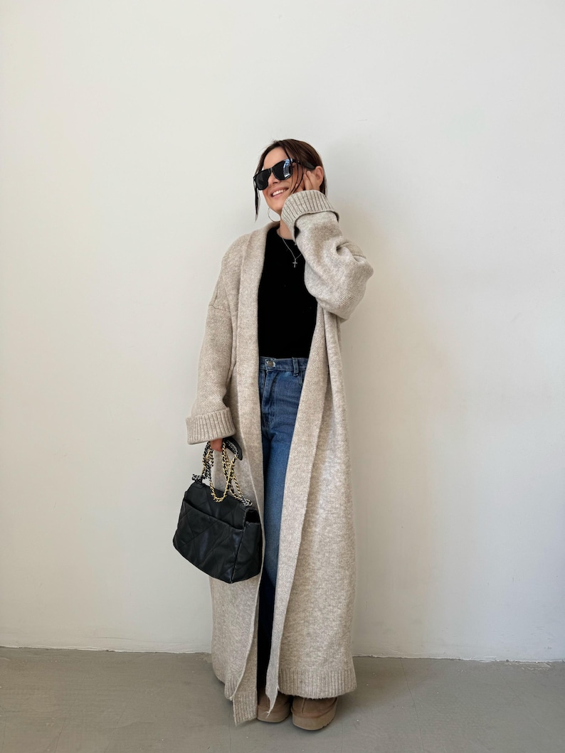 May include: A long, beige cardigan with a shawl collar and long sleeves. The cardigan is open in the front and is worn over a black top and blue jeans. The person is wearing black sunglasses and carrying a black handbag with a gold chain strap.