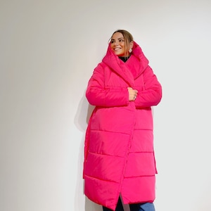 May include: A bright pink, long puffer coat with a wrap-style design. The coat features a large hood and a belt. The person is wearing blue jeans. The coat is a vibrant, eye-catching piece of outerwear.