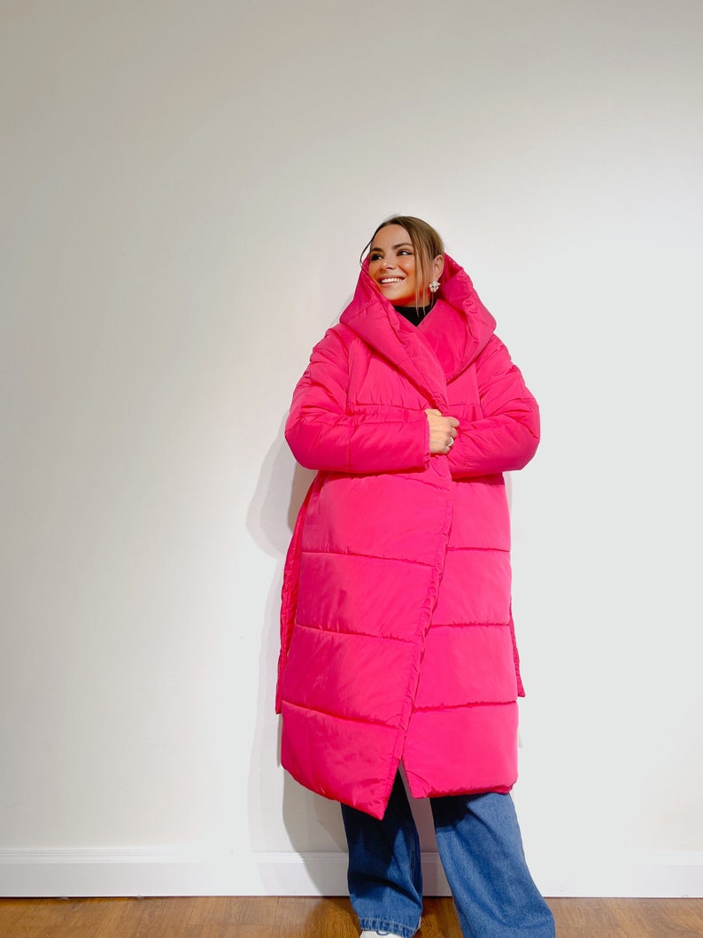 May include: A bright pink, long puffer coat with a wrap-style design. The coat features a large hood and a belt. The person is wearing blue jeans. The coat is a vibrant, eye-catching piece of outerwear.