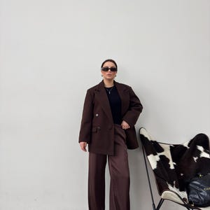 May include: A woman wearing a brown blazer and matching wide-leg trousers, with a black top and sunglasses. She is standing in front of a white wall with a cowhide chair in the background.