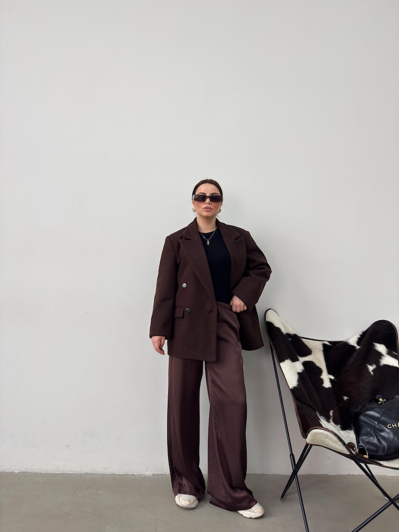 May include: A woman wearing a brown blazer and matching wide-leg trousers, with a black top and sunglasses. She is standing in front of a white wall with a cowhide chair in the background.