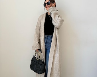 May include: A long, beige cardigan with a shawl collar and long sleeves. The cardigan is open in the front and is worn over a black top and blue jeans. The person is wearing black sunglasses and carrying a black handbag with a gold chain strap.
