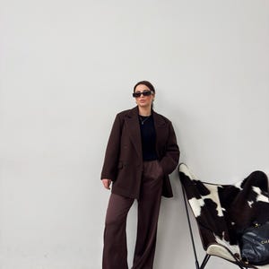 May include: A woman wearing a brown blazer and matching wide-leg trousers. She is wearing sunglasses and a black top. She is standing in front of a white wall with a white and black cowhide chair in the background.