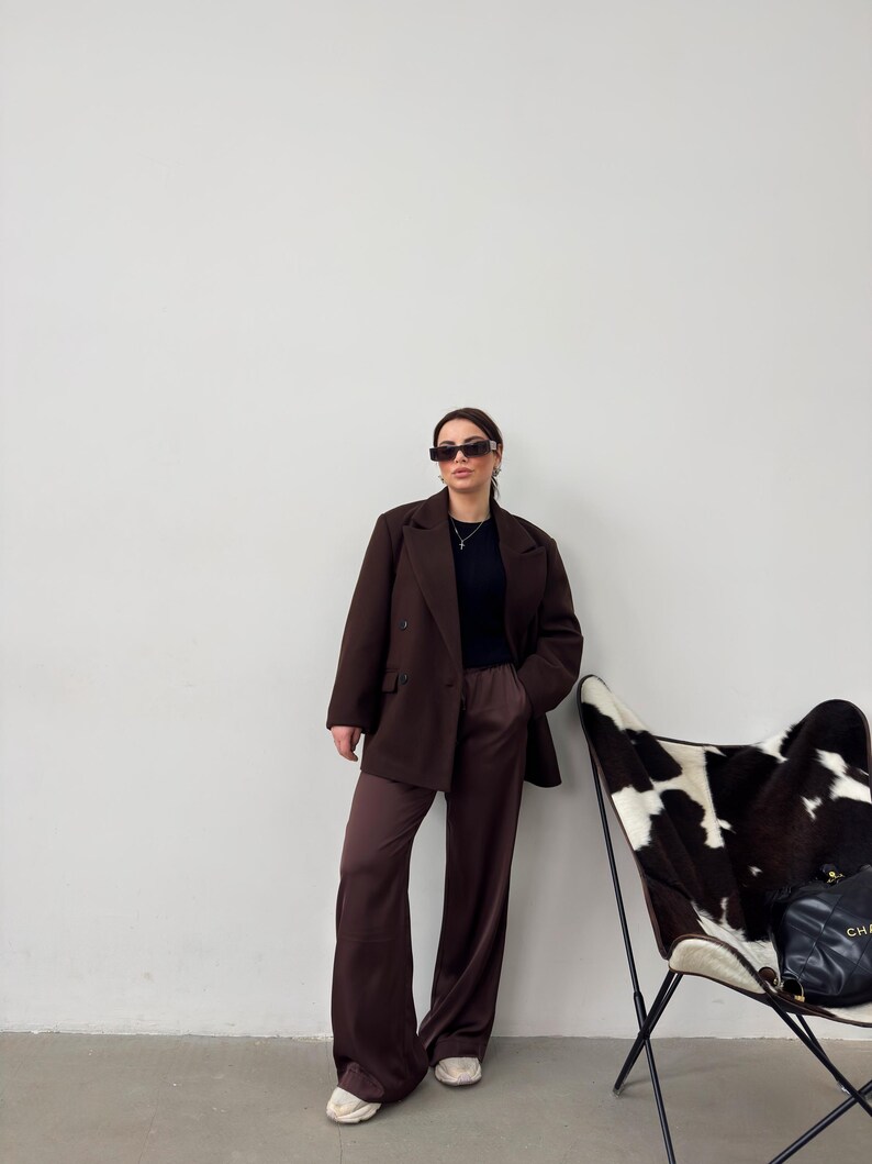 May include: A woman wearing a brown blazer and matching wide-leg trousers. She is wearing sunglasses and a black top. She is standing in front of a white wall with a white and black cowhide chair in the background.