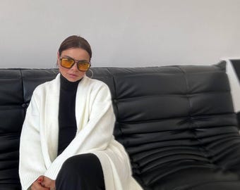Op de afbeelding: Een vrouw in een witte cardigan met een zwarte turtleneck en een zwarte broek zit op een zwarte leren bank. Ze draagt een gele zonnebril.